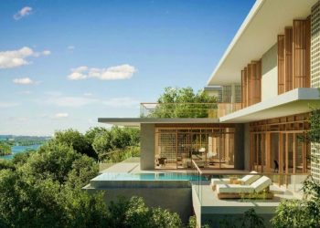 This stunning piece of India real estate features a modern house with large glass windows and wooden accents, surrounded by lush greenery. A terrace with lounge chairs and an infinity pool offers breathtaking views of trees and water under a clear blue sky, creating a serene escape. | MAKE1M