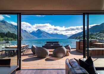 A spacious deck with modern wicker chairs and a dining table overlooks a scenic New Zealand lake surrounded by mountains under a blue sky, as viewed from inside a contemporary home through large glass doors. | MAKE1M