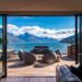 A spacious deck with modern wicker chairs and a dining table overlooks a scenic New Zealand lake surrounded by mountains under a blue sky, as viewed from inside a contemporary home through large glass doors. | MAKE1M