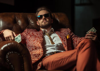 A man in a patterned suit and sunglasses lounges in a leather armchair, holding cash in both hands, exuding confidence and the success of high-ticket side hustles. | MAKE1M