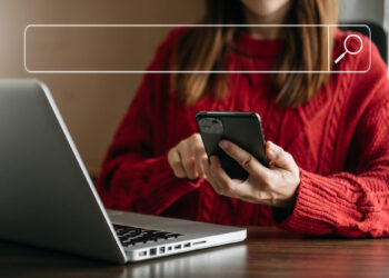 A person wearing a red sweater sits at a table with a laptop, holding a smartphone. A transparent search bar graphic appears above, symbolizing online searching with ClarityCheck under scrutiny for accurate results. | MAKE1M
