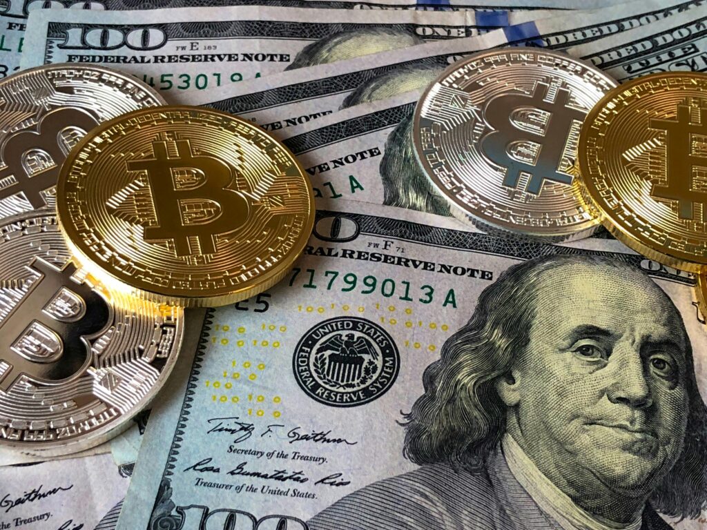 Several gold-colored Bitcoin tokens are placed on top of multiple U.S. one hundred dollar bills, highlighting the contrast between traditional cash and the digital asset revolution. | MAKE1M