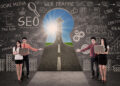 Four people with laptops stand in front of a chalkboard covered in SEO, web traffic, and social media doodles, pointing to a road leading to sunlight through a keyhole-shaped opening. | MAKE1M