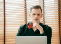 A man in a black shirt looks thoughtfully at a laptop while holding a credit card, sitting indoors with wooden blinds in the background. | MAKE1M