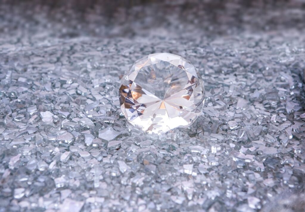 A large, round-cut diamond sits on a sparkling surface covered with small, clear, crushed crystals, reflecting light and creating a shimmering effect. | MAKE1M
