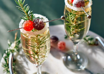 Two champagne glasses garnished with sugared cranberries and rosemary sprigs on a marble tray. Additional sugared cranberries and rosemary surround the glasses, creating a festive and elegant presentation. | MAKE1M