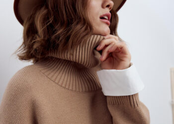 A woman with wavy brown hair wears a beige turtleneck sweater, a wide-brimmed brown hat, and a white shirt cuff, holding the collar of her sweater near her chin. The image is cropped to show her face in partial profile. | MAKE1M