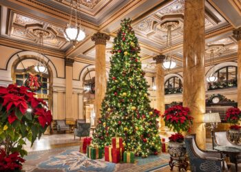 A grand, ornate hotel lobby decorated for Christmas with a large, lit Christmas tree surrounded by wrapped gifts, red poinsettias, marble columns, elegant chandeliers, and detailed ceiling moldings. | MAKE1M