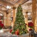 A grand, ornate hotel lobby decorated for Christmas with a large, lit Christmas tree surrounded by wrapped gifts, red poinsettias, marble columns, elegant chandeliers, and detailed ceiling moldings. | MAKE1M