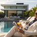 A man in a light suit and sunglasses relaxes on a lounge chair by a pool, reading a magazine and holding a drink, with a modern house and lush greenery in the background. | MAKE1M