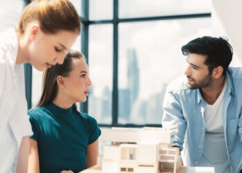 Three people are having a discussion in a bright office with large windows. Two women and one man are focused on a table with architectural models, with a cityscape visible in the background. | MAKE1M