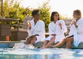 Three women in white robes sit by a pool, smiling and holding glasses of champagne. Two women have their feet in the water. Outdoor lounge chairs and greenery are in the background on a sunny day. | MAKE1M