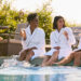 Three women in white robes sit by a pool, smiling and holding glasses of champagne. Two women have their feet in the water. Outdoor lounge chairs and greenery are in the background on a sunny day. | MAKE1M