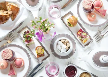 An elegant afternoon tea setting with teapots, teacups, assorted pastries, macarons, scones, finger sandwiches, sugar cubes, and a small vase of pink flowers, all arranged on a white tablecloth. | MAKE1M