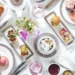 An elegant afternoon tea setting with teapots, teacups, assorted pastries, macarons, scones, finger sandwiches, sugar cubes, and a small vase of pink flowers, all arranged on a white tablecloth. | MAKE1M