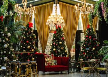 A festive room with gold drapes, chandeliers, and greenery, features several decorated Christmas trees with red bows and ornaments, a red velvet sofa, and gold tables and chairs. | MAKE1M