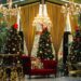 A festive room with gold drapes, chandeliers, and greenery, features several decorated Christmas trees with red bows and ornaments, a red velvet sofa, and gold tables and chairs. | MAKE1M
