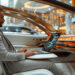 A man in a light grey suit works on a laptop inside a luxurious, futuristic car with a sleek high-tech interior and ambient orange lighting, parked in a modern, glass-walled environment. | MAKE1M