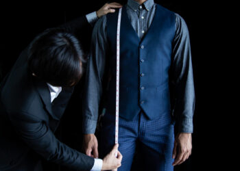A tailor in a black suit measures the sleeve length of a man wearing a navy vest, gray shirt, and blue checkered pants, using a measuring tape against a black background. | MAKE1M
