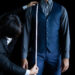 A tailor in a black suit measures the sleeve length of a man wearing a navy vest, gray shirt, and blue checkered pants, using a measuring tape against a black background. | MAKE1M