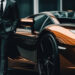 A person in a dark suit stands beside a shiny orange sports car, possibly a luxury model, in an urban setting with modern glass buildings in the background. | MAKE1M