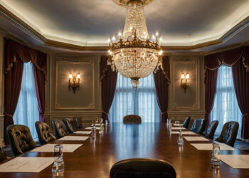 Elegant boardroom with a large wooden table, leather chairs, chandelier, ornate wall sconces, and tall windows draped with burgundy curtains. Notepads and glasses of water are set at each seat. | MAKE1M