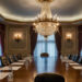 Elegant boardroom with a large wooden table, leather chairs, chandelier, ornate wall sconces, and tall windows draped with burgundy curtains. Notepads and glasses of water are set at each seat. | MAKE1M