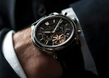 A close-up of a person wearing a luxury black and silver mechanical wristwatch with visible gears and multiple dials, dressed in a dark suit. | MAKE1M