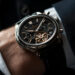 A close-up of a person wearing a luxury black and silver mechanical wristwatch with visible gears and multiple dials, dressed in a dark suit. | MAKE1M