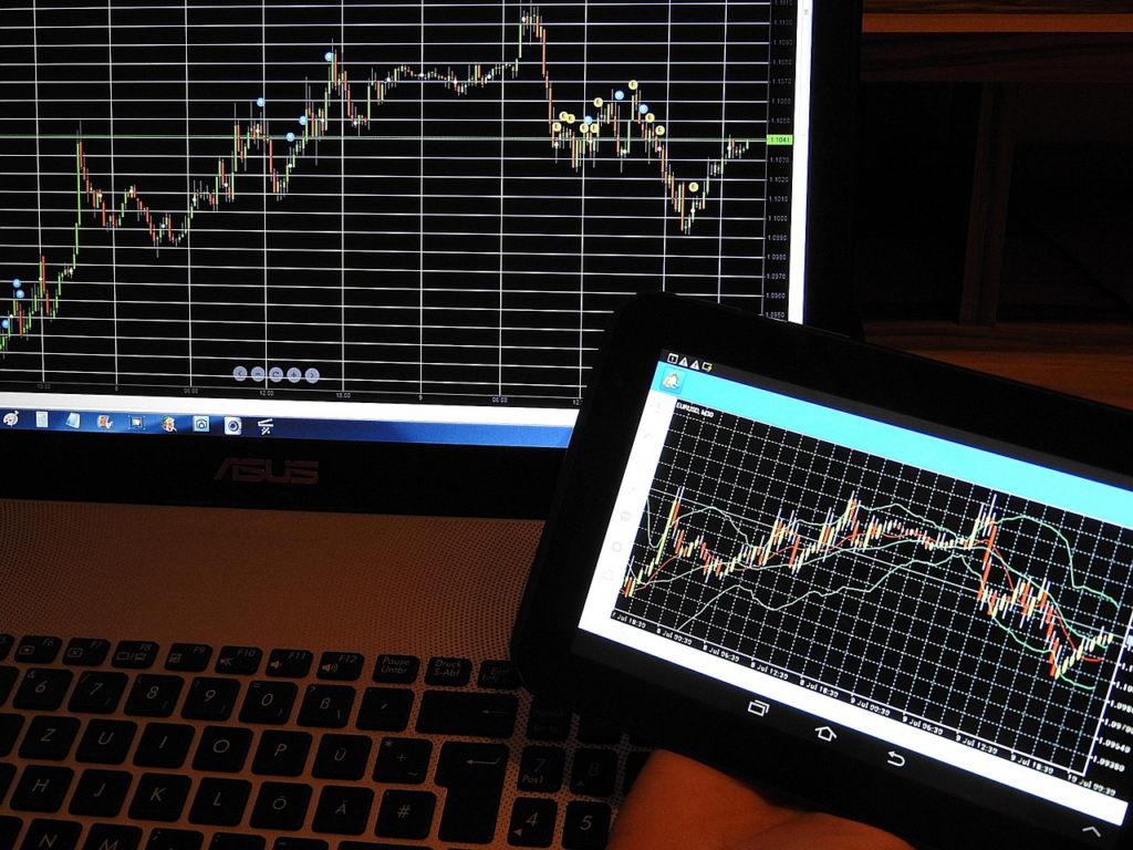 A person holds a tablet displaying a stock or financial chart in front of a laptop with a similar chart on its screen. Both charts show fluctuating lines and data points on a grid background. | MAKE1M