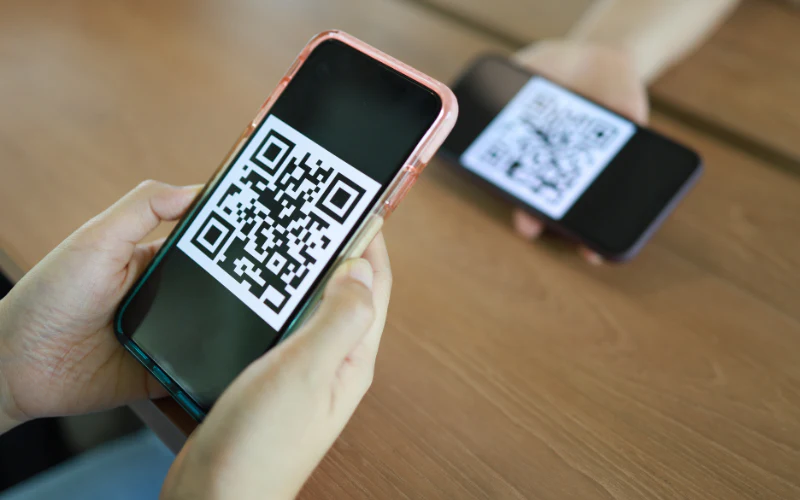 Two people holding smartphones displaying QR codes on their screens over a wooden table, possibly for scanning or digital payment purposes. | MAKE1M