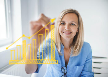 A woman in a blue shirt smiles while drawing an upward-trending yellow bar and line graph on a transparent screen in a bright office setting. | MAKE1M
