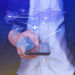 A person holding a smartphone with a digital overlay of a world map and airplane icons connected by lines, symbolizing global travel or online flight booking. | MAKE1M