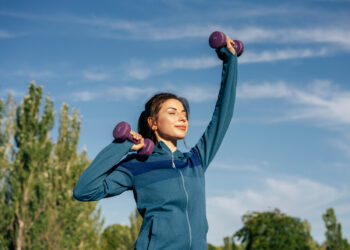 A woman in a blue athletic jacket lifts purple dumbbells outdoors, smiling and closing her eyes under a clear blue sky with trees in the background. | MAKE1M