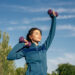 A woman in a blue athletic jacket lifts purple dumbbells outdoors, smiling and closing her eyes under a clear blue sky with trees in the background. | MAKE1M