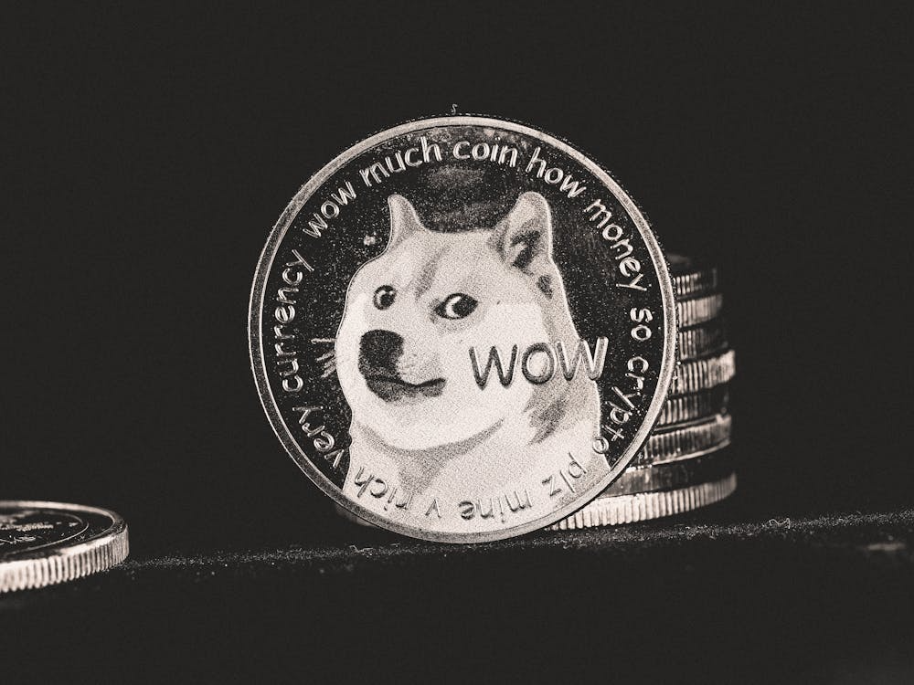 A close-up of a Dogecoin, a cryptocurrency coin featuring the Shiba Inu dog meme, with the words "wow much coin how money so crypto" around the edge. Several coins are stacked in the background. | MAKE1M