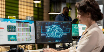 A woman works at a desk with two monitors; one displays AI statistics and data, while the other shows a digital brain graphic, suggesting she is analyzing artificial intelligence or machine learning information. | MAKE1M
