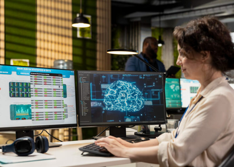 A woman works at a desk with two monitors; one displays AI statistics and data, while the other shows a digital brain graphic, suggesting she is analyzing artificial intelligence or machine learning information. | MAKE1M