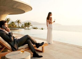 A man in a suit sits on a lounge chair by a pool, talking on the phone, while a woman in a white dress stands nearby holding a drink, overlooking the ocean at sunset. | MAKE1M