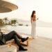 A man in a suit sits on a lounge chair by a pool, talking on the phone, while a woman in a white dress stands nearby holding a drink, overlooking the ocean at sunset. | MAKE1M