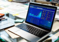 A laptop on a glass desk displays stock market charts and data. Papers, a smartphone, sticky notes, and a water glass are also visible, suggesting a busy financial or office workspace. | MAKE1M