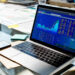 A laptop on a glass desk displays stock market charts and data. Papers, a smartphone, sticky notes, and a water glass are also visible, suggesting a busy financial or office workspace. | MAKE1M