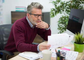 A man with gray hair and glasses sits at a desk, looking thoughtfully at a paper with charts and graphs—perhaps reviewing data related to wealth. A computer, potted plant, mug, and calendar are on the desk in a modern office setting. | MAKE1M