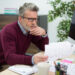 A man with gray hair and glasses sits at a desk, looking thoughtfully at a paper with charts and graphs—perhaps reviewing data related to wealth. A computer, potted plant, mug, and calendar are on the desk in a modern office setting. | MAKE1M